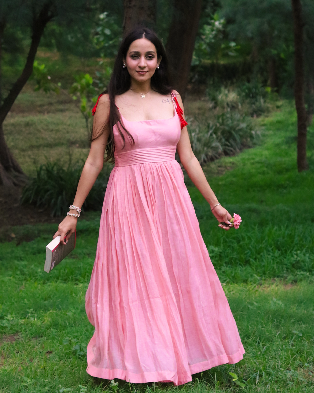 A woman wearing a flowing pink sleeveless maxi dress with red shoulder ties stands outdoors on a grassy path surrounded by trees. She is holding a small book or clutch in one hand and a small pink flower in the other.