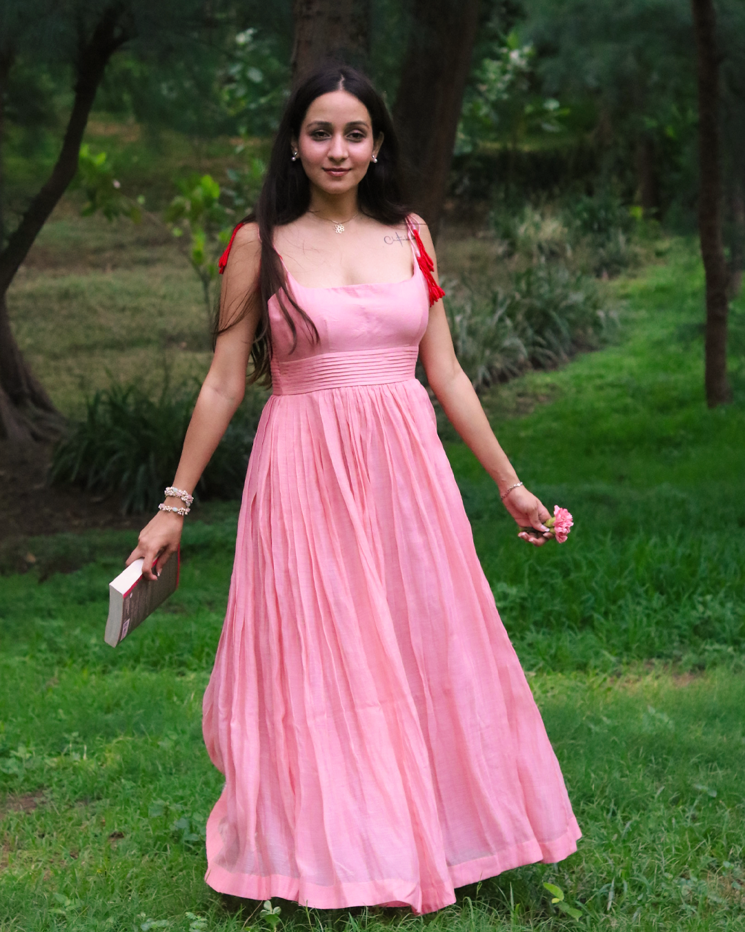 A woman wearing a flowing pink sleeveless maxi dress with red shoulder ties stands outdoors on a grassy path surrounded by trees. She is holding a small book or clutch in one hand and a small pink flower in the other.