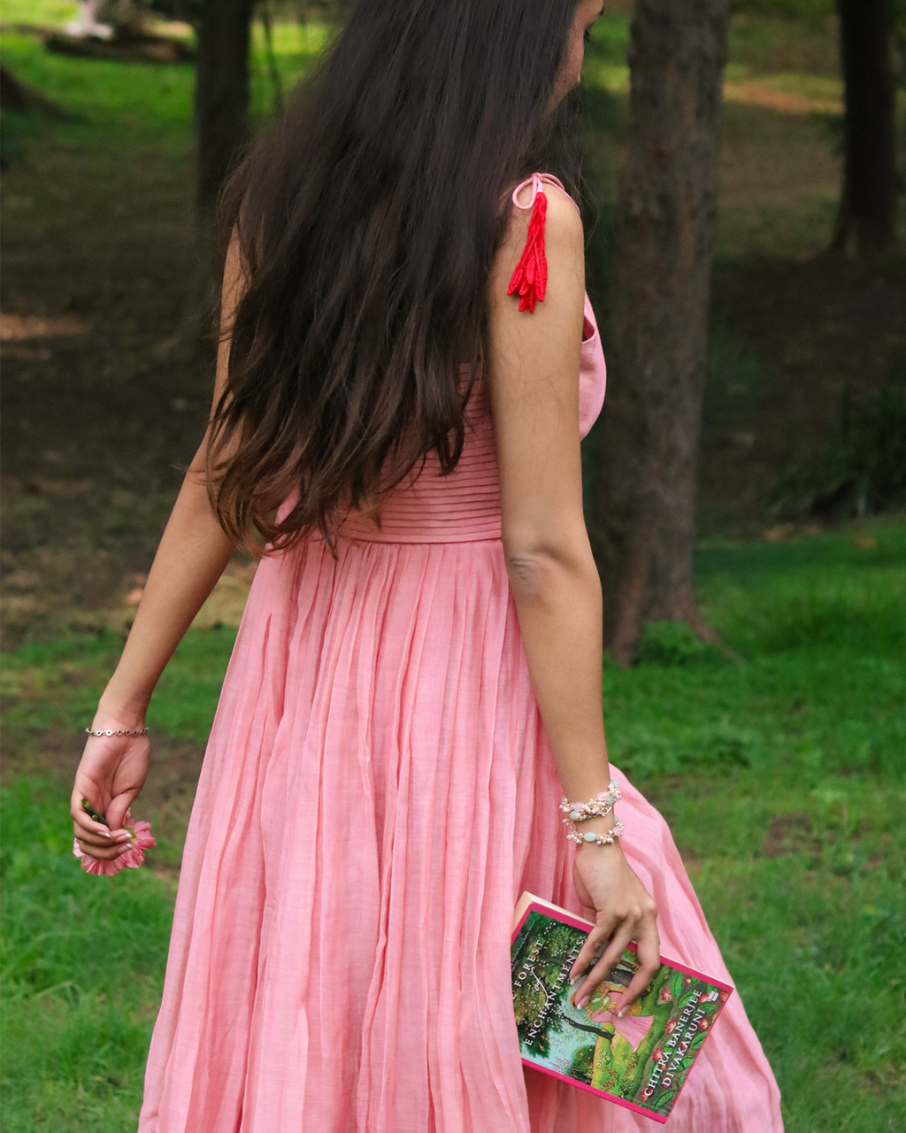 A woman wearing a flowing pink sleeveless maxi dress with red tassel shoulder ties, photographed outdoors in a park among trees and grass. She is holding a colorful book.