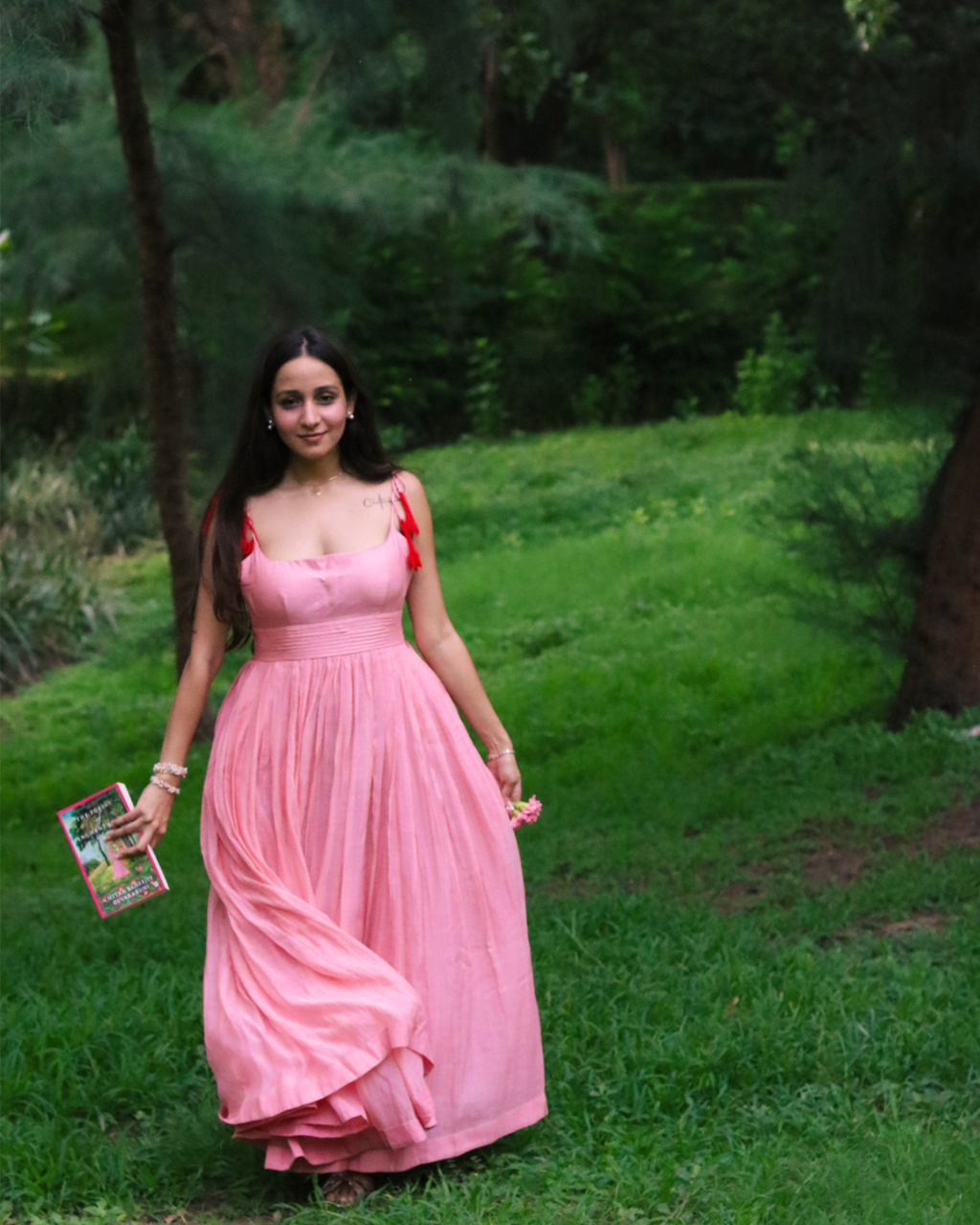 A woman wearing a pink sleeveless maxi dress with red shoulder ties stands on a grassy park path, holding a magazine.